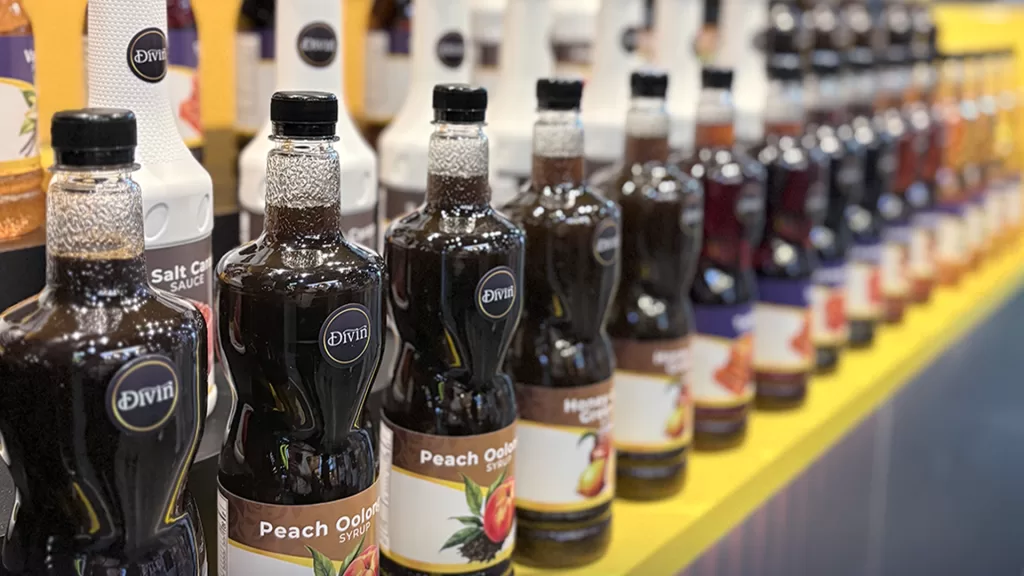 A close-up of Divin's product lineup displayed at WOFEX 2025, featuring rows of naturally flavored syrup bottles including Peach Oolong and Salted Caramel, neatly arranged on a vibrant yellow shelf.